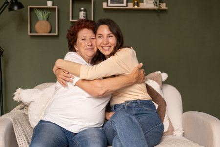 Portrait of a modern happy grandmother and granddaughter sitting in the living room during the day on the sofa, care hugging and looking awayの写真素材
