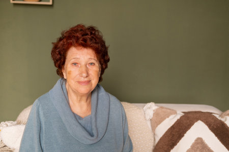 Portrait of an elderly woman with blue eyes and red hair looks at the camera in a room sitting on a...
