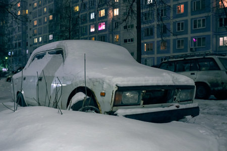 Old car covered with snow near a multi-storey building at nightの写真素材
