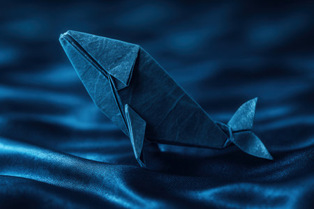 Blue paper origami whale figure swimming on wavy blue silk fabric background resembling ocean waterの素材