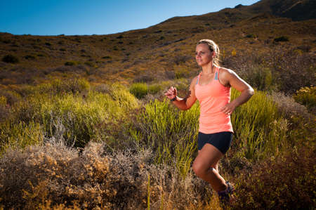 Vibrant image of an athletic looking blonde female trail runner running along an unseen mountain path at sunsetの写真素材