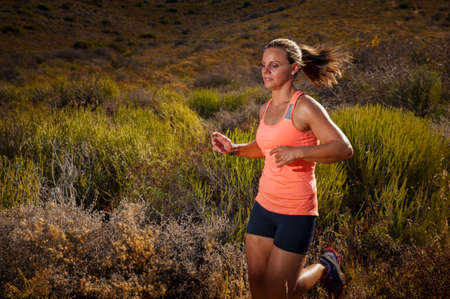 Vibrant image of an athletic looking blonde female trail runner running along an unseen mountain path at sunsetの写真素材