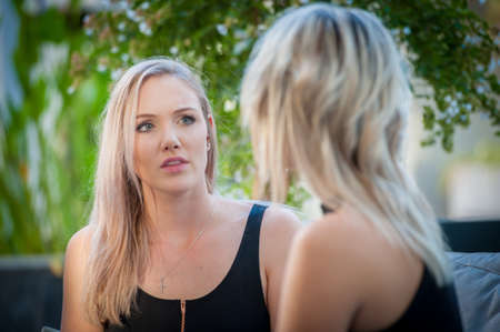 two pretty blonde sisters sat on a sofa outside enjoying a coffeeの写真素材