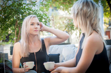 two pretty blonde sisters sat on a sofa outside enjoying a coffeeの写真素材