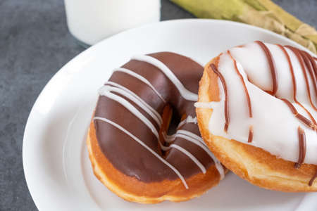 two white and brown iced ring doughnuts on a white plate with milk, a napkin and slate background