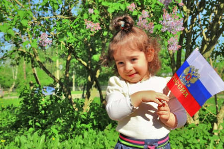 Little baby girl stands with flag of Russia in front of lilac bush on sunny day. Russian translation on flag: Russiaの写真素材