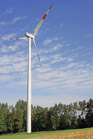 An image of a singular wind turbinestanding alone against a forest with a blue sky and clouds behindの写真素材