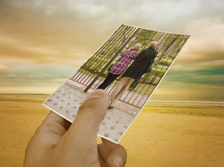 Concept creative collage with a hand holding a photo of a couple of mature women and a nice isolated beach in the backgroundの写真素材