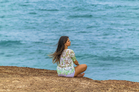 PIPA, BRAZIL, JANUARY - 2016 - Young attractive woman sitting at edge of rocky hill with the ocean at the background in Pipa, Brazilのeditorial素材