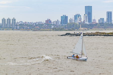 MONTEVIDEO, URUGUAY, JANUARY -2016 - Small sailboat with two persons sailing at river with Montevideo skyline at backgroundのeditorial素材