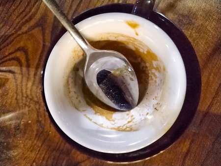 Overhead view of an empty coffee cup with a spoon resting inside. The cup has remnants of coffee stains clinging to the sides, and it sits on a saucer atop a wooden table.の写真素材
