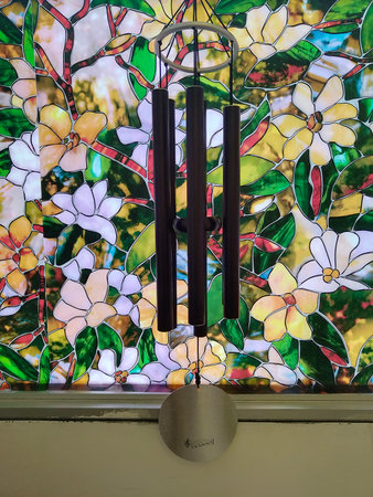 A black wind chime hanging in front of a vibrant stained glass window with floral designs.の写真素材