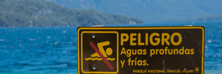 Wooden signpost with "danger, deep and cold waters" spanish text at one of the beaches of bariloche, nahuel huapi national park, rio negro province, argentina.の写真素材