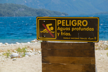 Wooden signpost with "danger, deep and cold waters" spanish text at one of the beaches of bariloche, nahuel huapi national park, rio negro province, argentina.の写真素材