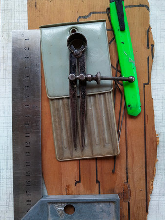 Collection of tools on a wooden surface with drawn outlines. The items include a metal ruler, an old rusty compass, and a green utility cutter.の写真素材