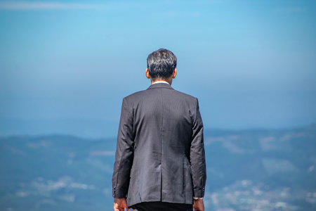 Rear medium shot of a man in a black suit standing still, facing an open mountain landscapeの写真素材