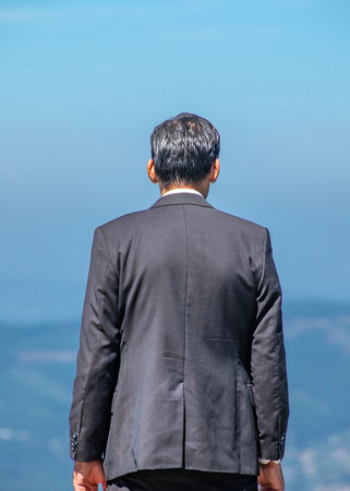 Rear medium shot of a man in a black suit standing still, facing an open mountain landscapeの写真素材