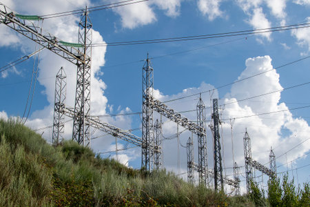 A wide, low-angle shot of a large electrical power substation situated on a green hill under a blue...