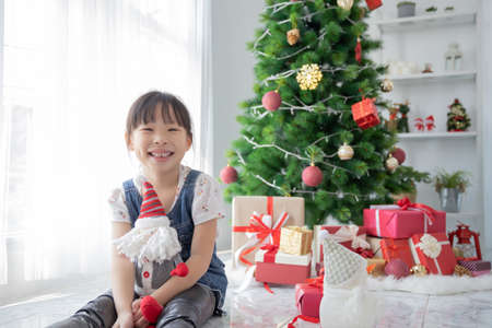 Cute Asian little girl holding Santa Claus doll in room with decorated with Christmas treeの写真素材