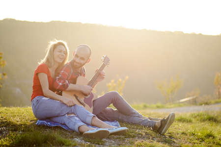 Young couple in love sitting on the park  while these young guitar playing guitar in sunset time.の写真素材