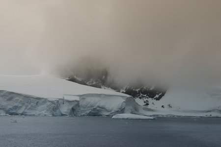 This image features a serene Antarctic landscape where icebergs meet the mist-covered mountains. The subdued colors and the vast, quiet expanse of water convey a sense of tranquility and the untouched beauty of the polar region.の写真素材