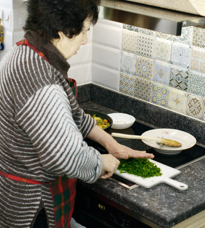 An elderly woman finely chopping fresh parsley on a cutting board, with a pot of vegetable stew simmering beside herの写真素材