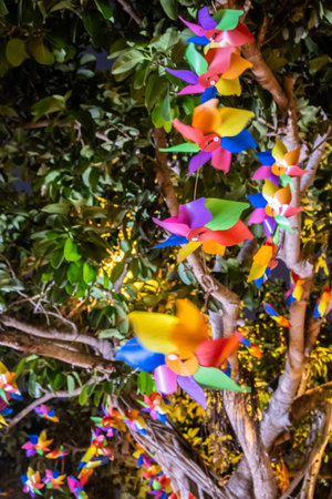 A tree trunk is decorated with colorful pinwheels, each with four blades in a variety of hues. The pinwheels are strung together and hang from the branches, creating a cheerful and festive atmosphere.の写真素材