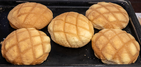 Five golden-brown melon pan breads, with a distinctive cross-hatch pattern, sit on a dark baking...