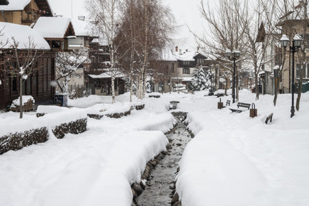 A picturesque pedestrian walkway in the mountain town of Bansko, Bulgaria covered in fresh snow, with a central creek, traditional houses, and bare trees on a cold winter day.の写真素材