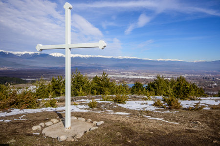 A large white Christian cross on a snowy hilltop overlooks a valley and Bansko, with the snow-capped Pirin mountains in the distance on a clear day.の写真素材