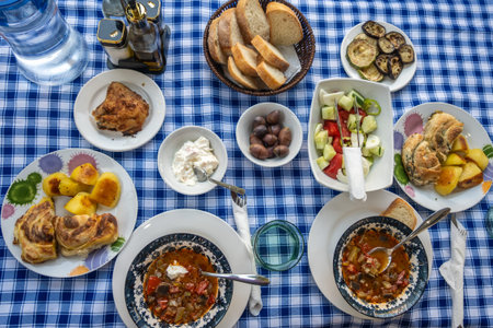 A rich assortment of traditional dishes including stews, roasted chicken, potatoes, bread, olives, salad, and a creamy dip, arranged on a vibrant checkered tablecloth.