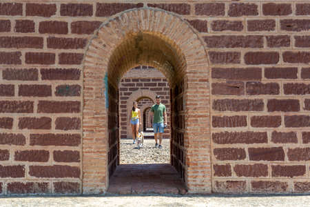 A caucasian young couple and their dog walking through an arched bridge on a sunny morning day