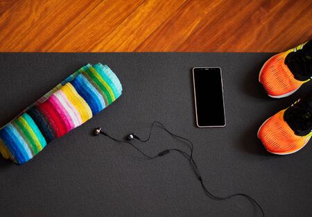 sports mat with mobile phone, headphones, orange sneakers and colorful lgtb towel on gymnasium floor or wooden house. Sport, healthy life and technology.の写真素材