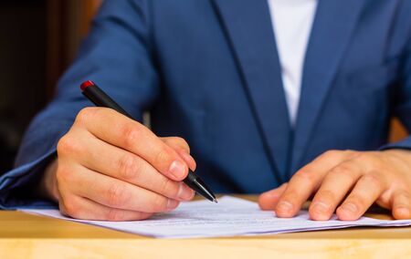 Close-up of the hands of a well-dressed man in a blue suit and white shirt signing a document on a...