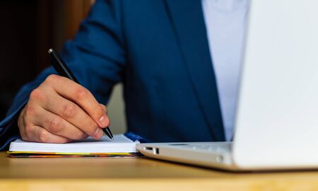Close-up of the hands of a well-dressed man in a blue suit and a white shirt writing in an agenda or a notebook, with a laptop on a deskの写真素材