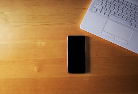 Black mobile phone and computer with white keyboard on top of a wooden office table. With space in the imageの写真素材