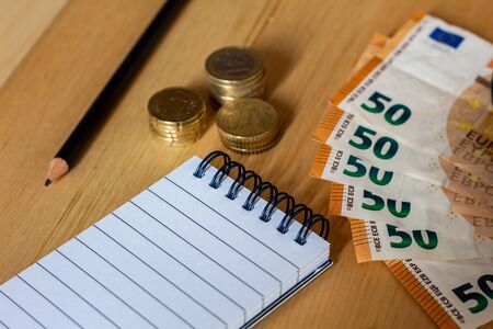 Notebook or notepad and pencil, surrounded by coins and bills, on a wooden office tableの写真素材