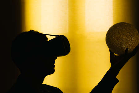 Silhouette of a young man with virtual reality glasses, VR, and a glowing ball in his hand. Hologram concept .Yellow background with copy spaceの写真素材