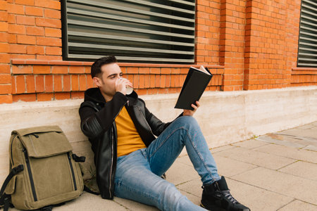 Handsome young man drinking a coffee and reading a book in the street, taking advantage of the good spring or autumn weather. with copy space.の写真素材