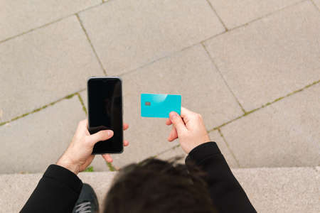 Young man holding a credit card and using his smartphone for online purchases and payments makes a...