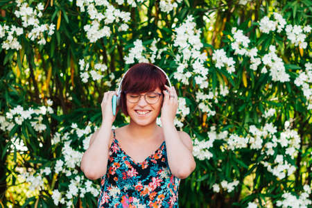 Young redhead woman listening to music with her headphones and mobile phone outdoors with the park...