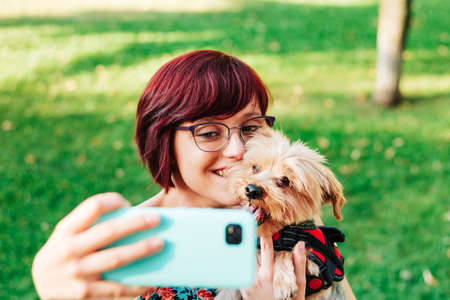 A person, young woman taking a photo or a selfie with her dog smiling and happy, with her mobile phone outdoors.の写真素材
