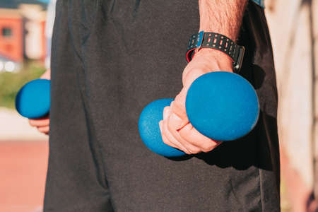 Close up shot of the hands of a sportsman holding weights with a smart whatch on his wrist while training outdoors. Sports and gym equipment concept.の写真素材