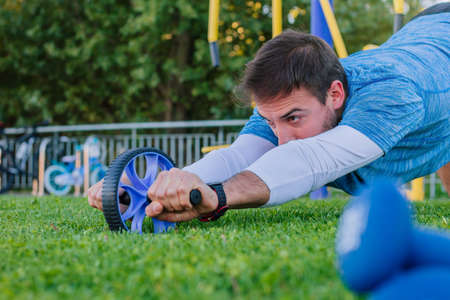 Young strong sports man training his abdomen muscles with a wheel outdoors in the natural park location.の写真素材