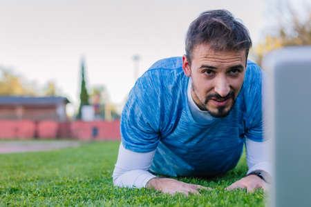 Athlete man doing an abdominal plank looking at his laptop while doing an online training and doing a sports course with a personal trainer.の写真素材