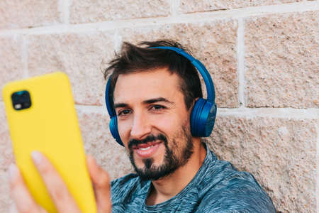 Young adult man dressed in sportswear using his yellow smartphone to take a selfie photo while listening to music with his headphones, resting from training.の写真素材