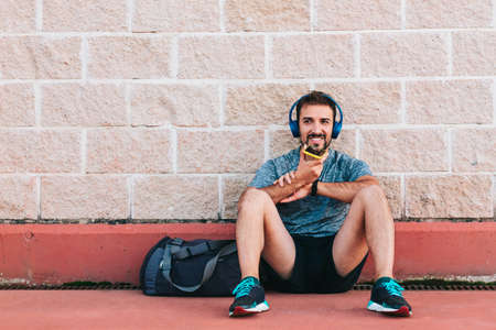 Young adult man dressed in sportswear and sports equipment, with his blue headphones and his yellow smartphone sitting on a wall outdoors with copy space.の写真素材