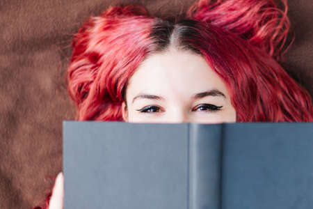 Young Caucasian woman, with white skin and red hair, smiling, amused and happy, while covering her face with a book while reading outdoors.の写真素材