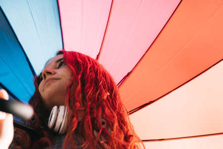 Young redhead 19s woman, with headphones and rainbow umbrella in the rain.の写真素材
