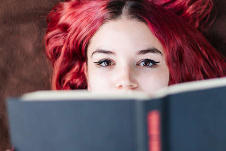 Young redhead teenage student, with white skin and freckles, relaxed reading a book in the park outdoors, enjoying reading looking at camera.の写真素材